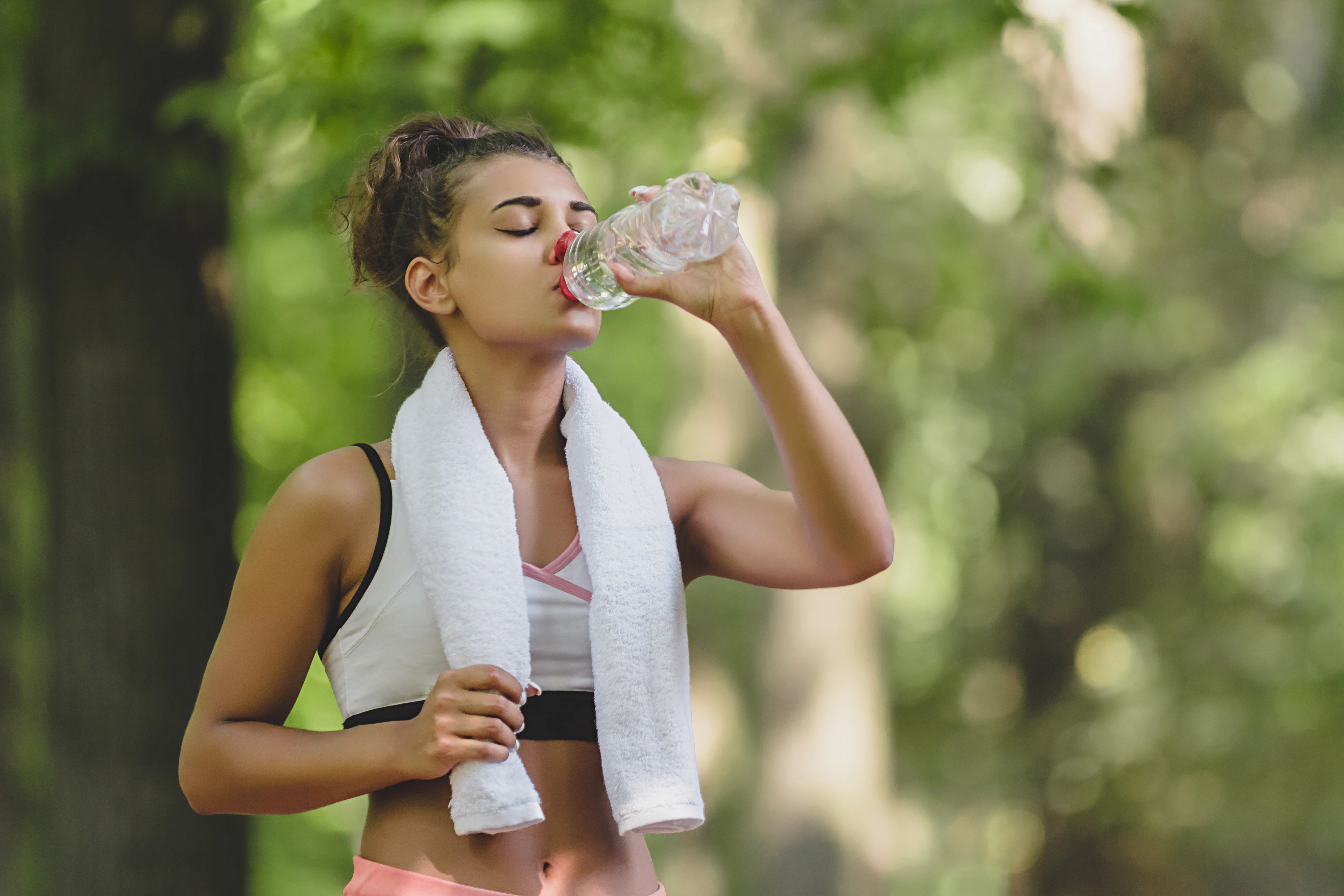 Beautiful young woman drinking water and cooling off after her running training at the park